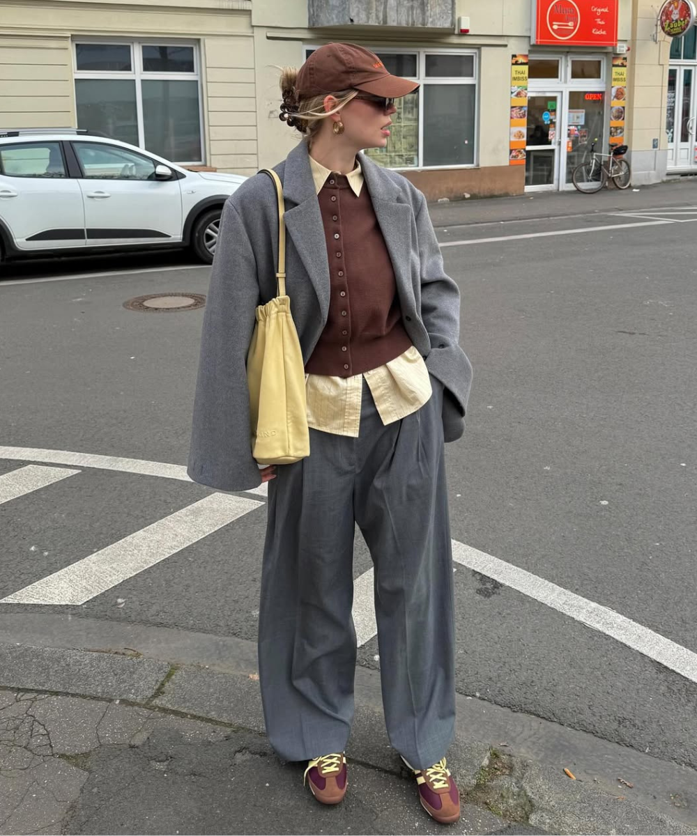 Pessoa usando casaco cinza sobre camisa e cardigã marrons, com calça larga cinza. Bolsa amarela e boné combinam na alfaiataria.