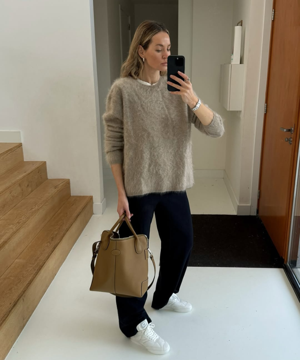 Pessoa com suéter de tricô bege, calça escura e tênis brancos, segurando bolsa marrom, em look casual.