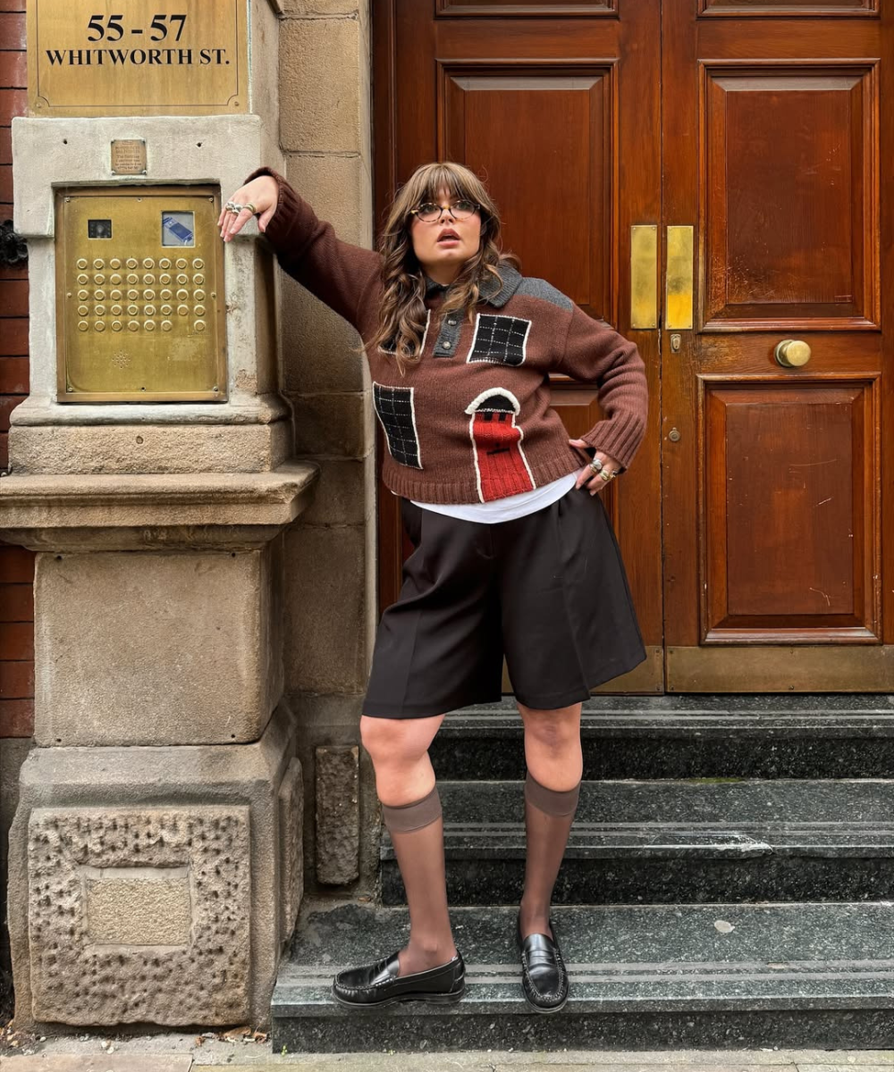 Pessoa com suéter de tricô estampado e shorts de alfaiataria marrom, em frente a uma porta de madeira.