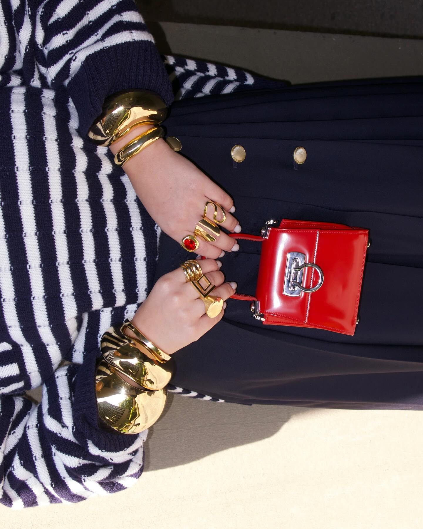 Suéter listrado, saia com botões dourados, bolsa vermelha, pulseiras volumosas, anéis modernos.