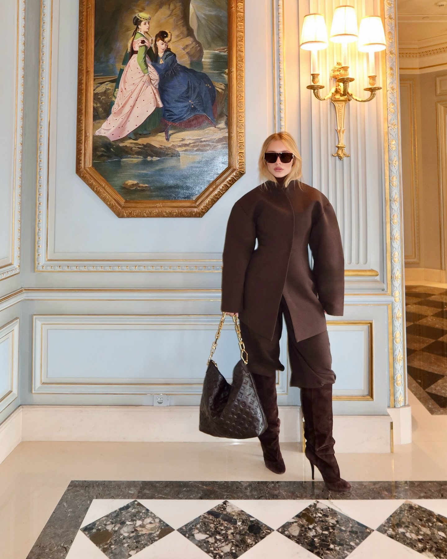 Pessoa em roupa elegante marrom, óculos escuros, bolsa, em interior sofisticado com quadro de mulheres ao fundo.