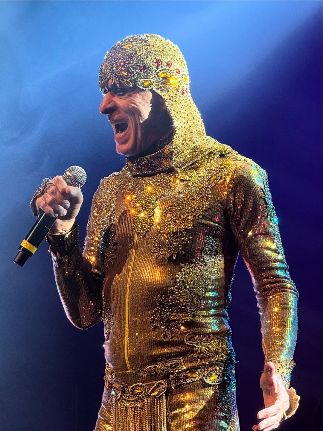 Cantor em um traje dourado brilhante, segurando um microfone e cantando no palco.