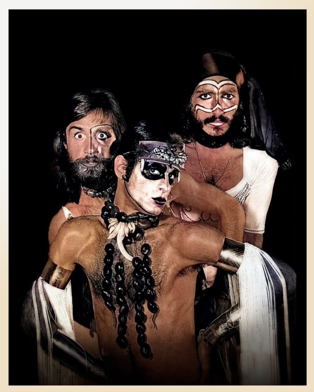 Três pessoas com maquiagem artística e figurinos coloridos. Elementos performáticos que remetem a Ney Matogrosso.