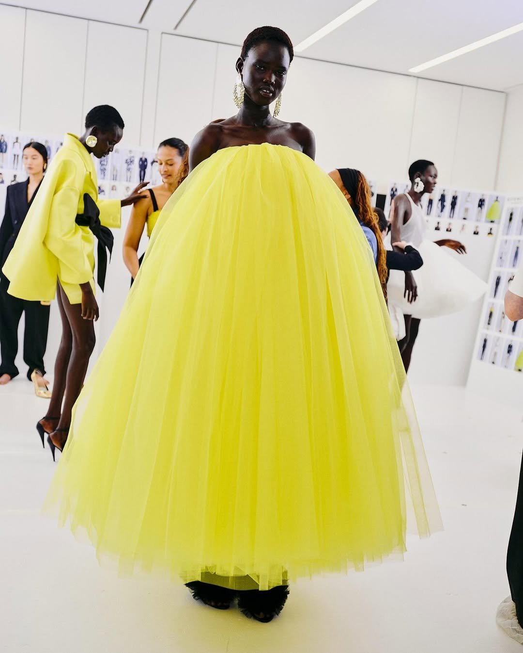 Modelo usa vestido volumoso amarelo em tons neon, em uma sala com pessoas ao fundo.