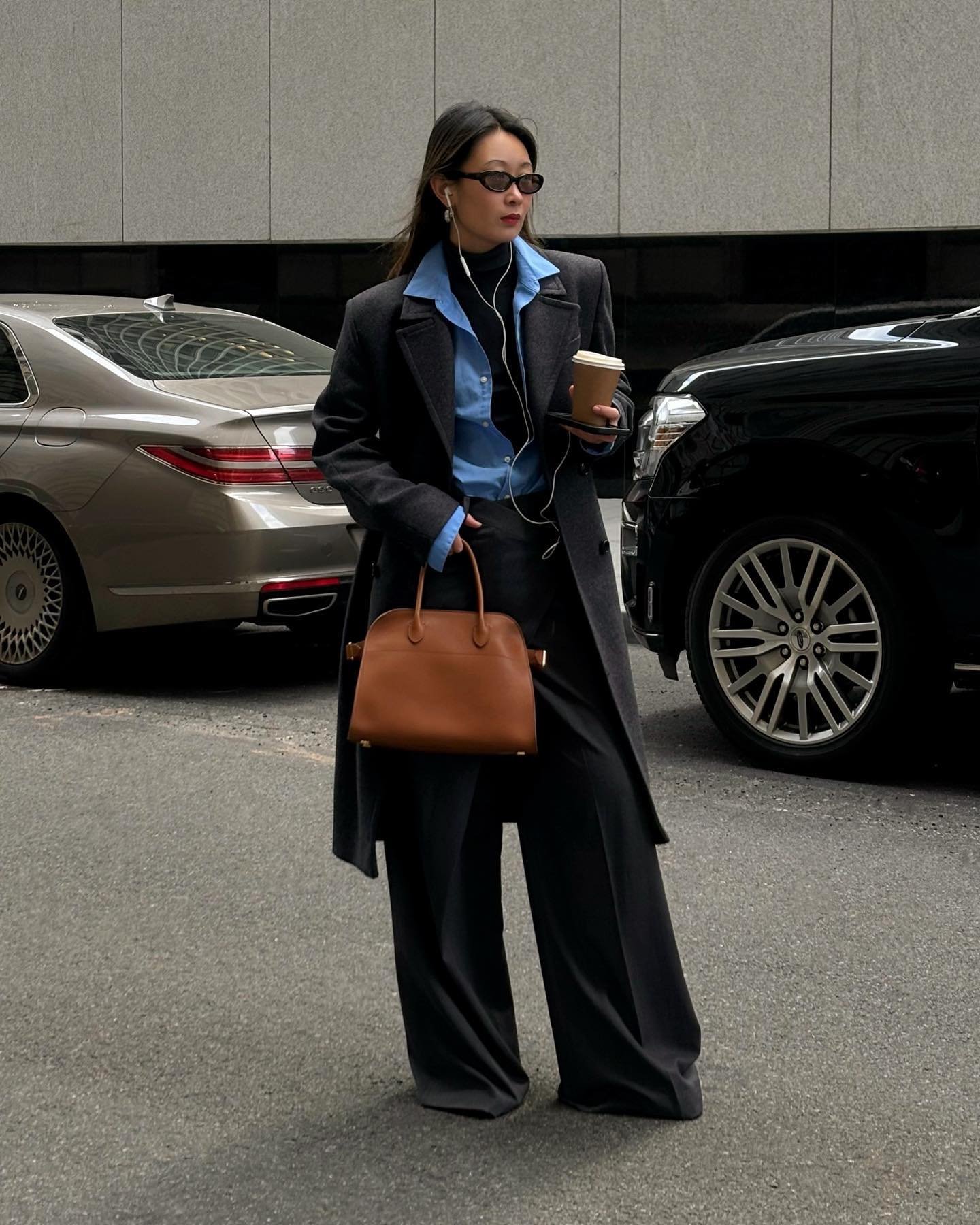 Pessoa com casaco escuro, blusa azul, bolsa marrom e copo na mão, oferecendo ideias de looks de frio.