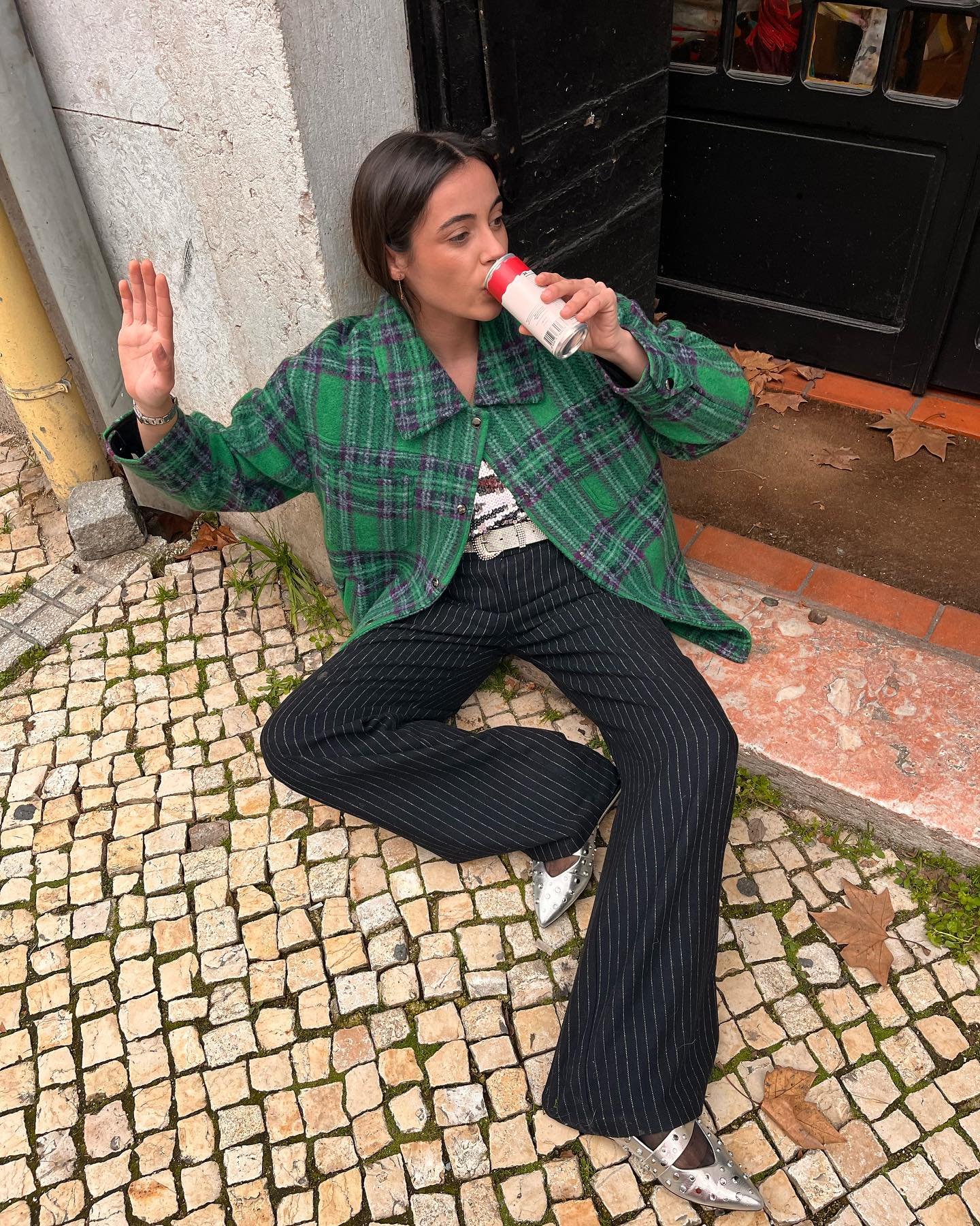 Pessoa sentada no chão, usando casaco xadrez verde sobre blusa de frio e calças listradas. Segurando bebida.