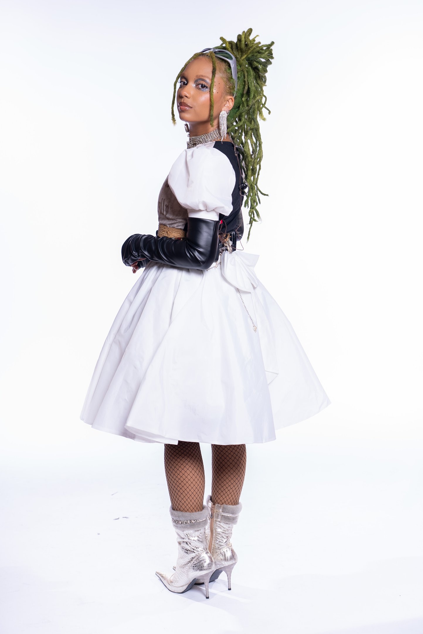 Nátaly Neri usando vestido branco com luvas pretas longas, cabelo verde preso, e botas prateadas.