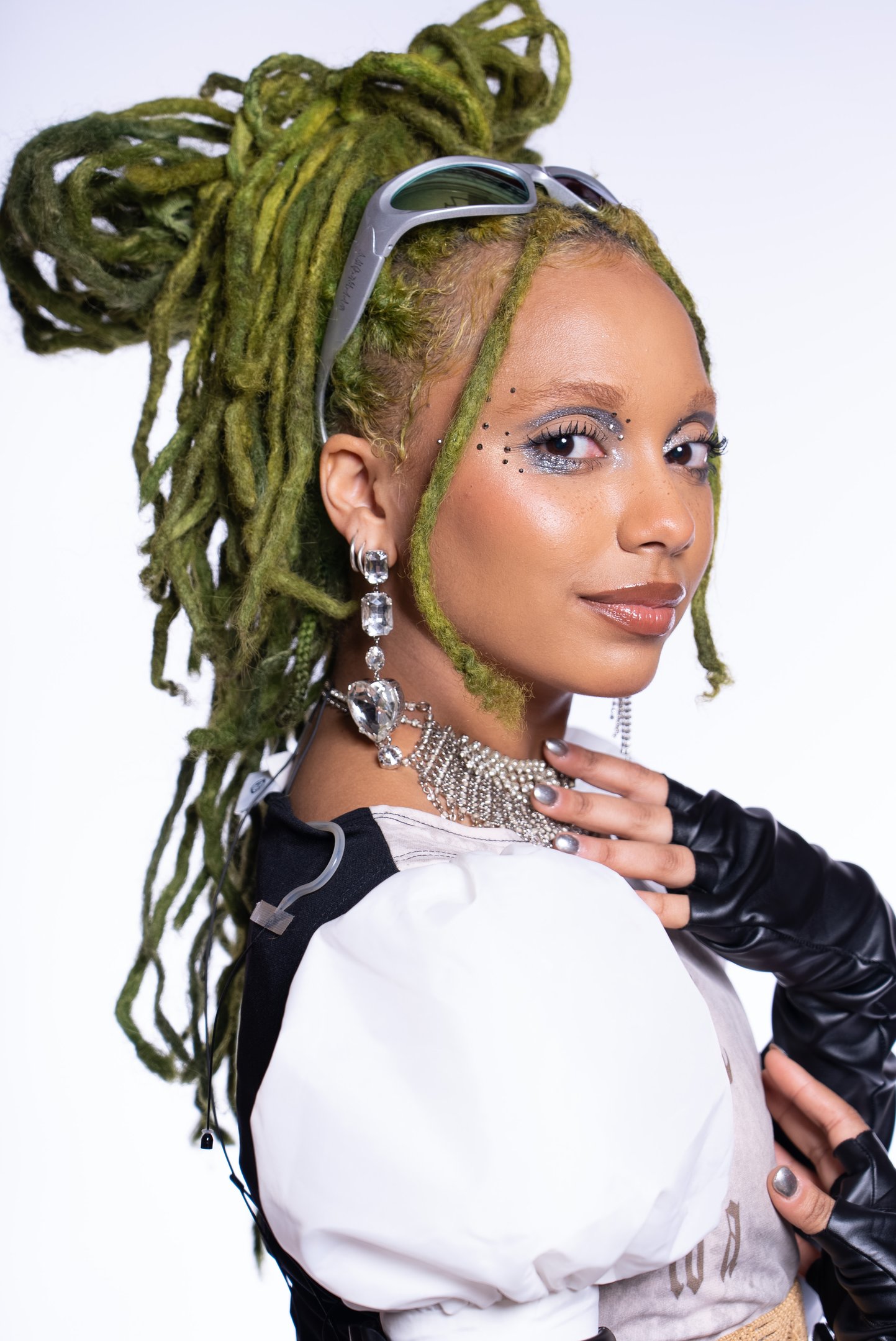 Nátaly Neri com dreadlocks verdes, óculos cinzas no topo da cabeça, maquiagem brilhante e acessórios prateados sofisticados.