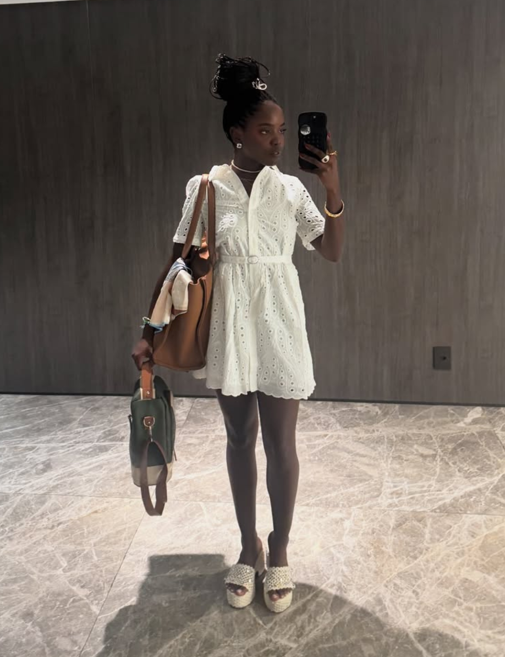 Pessoa com vestido branco elegante, segurando uma bolsa grande e lancheiras estilosas em um ambiente fechado.