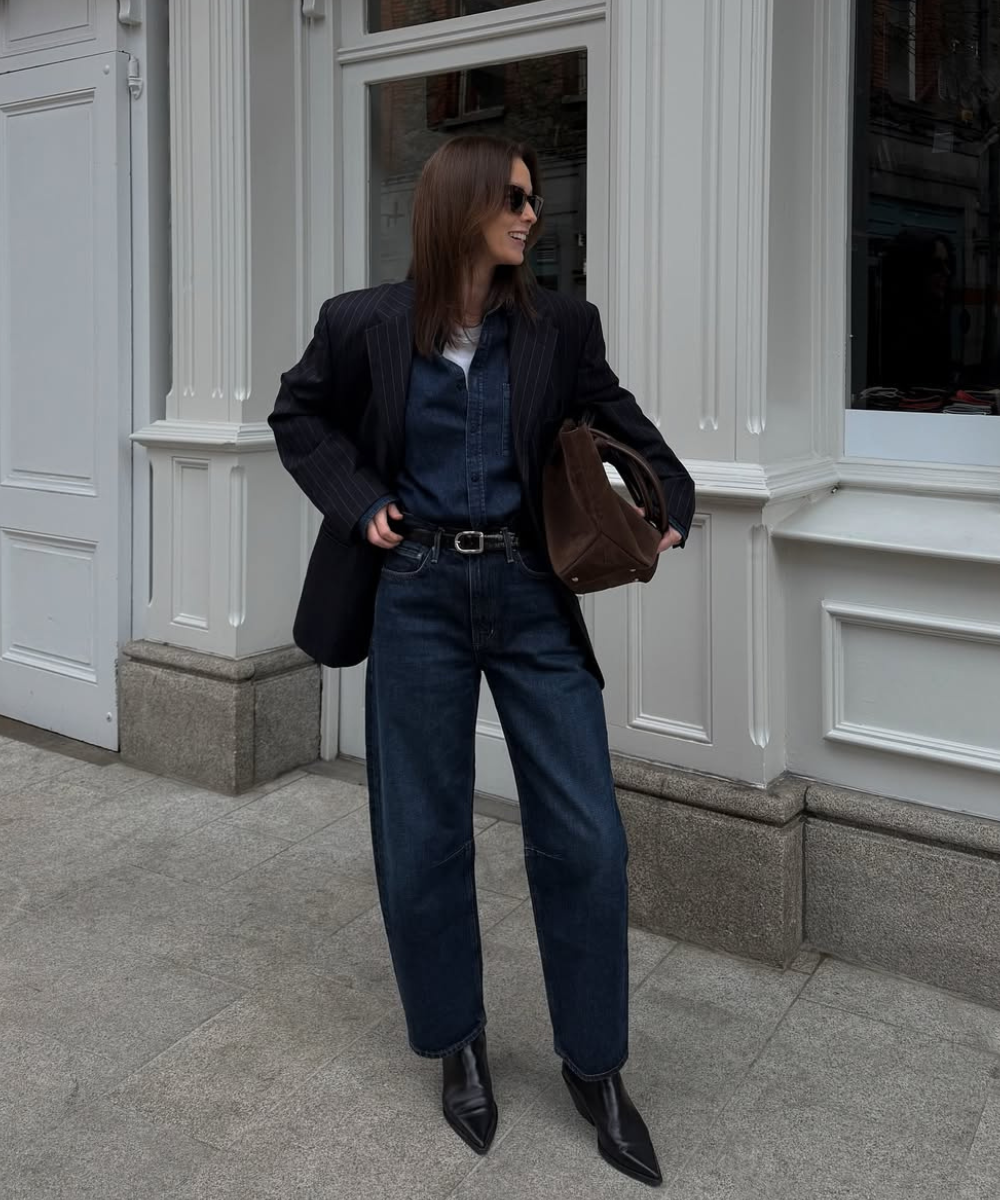 Pessoa usando blazer escuro sobre macacão jeans, em ambiente urbano, exemplificando como usar alfaiataria no dia a dia.