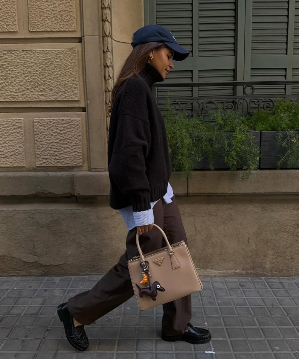 Pessoa caminha com estilo casual e calças de alfaiataria marrom, segurando uma bolsa bege.