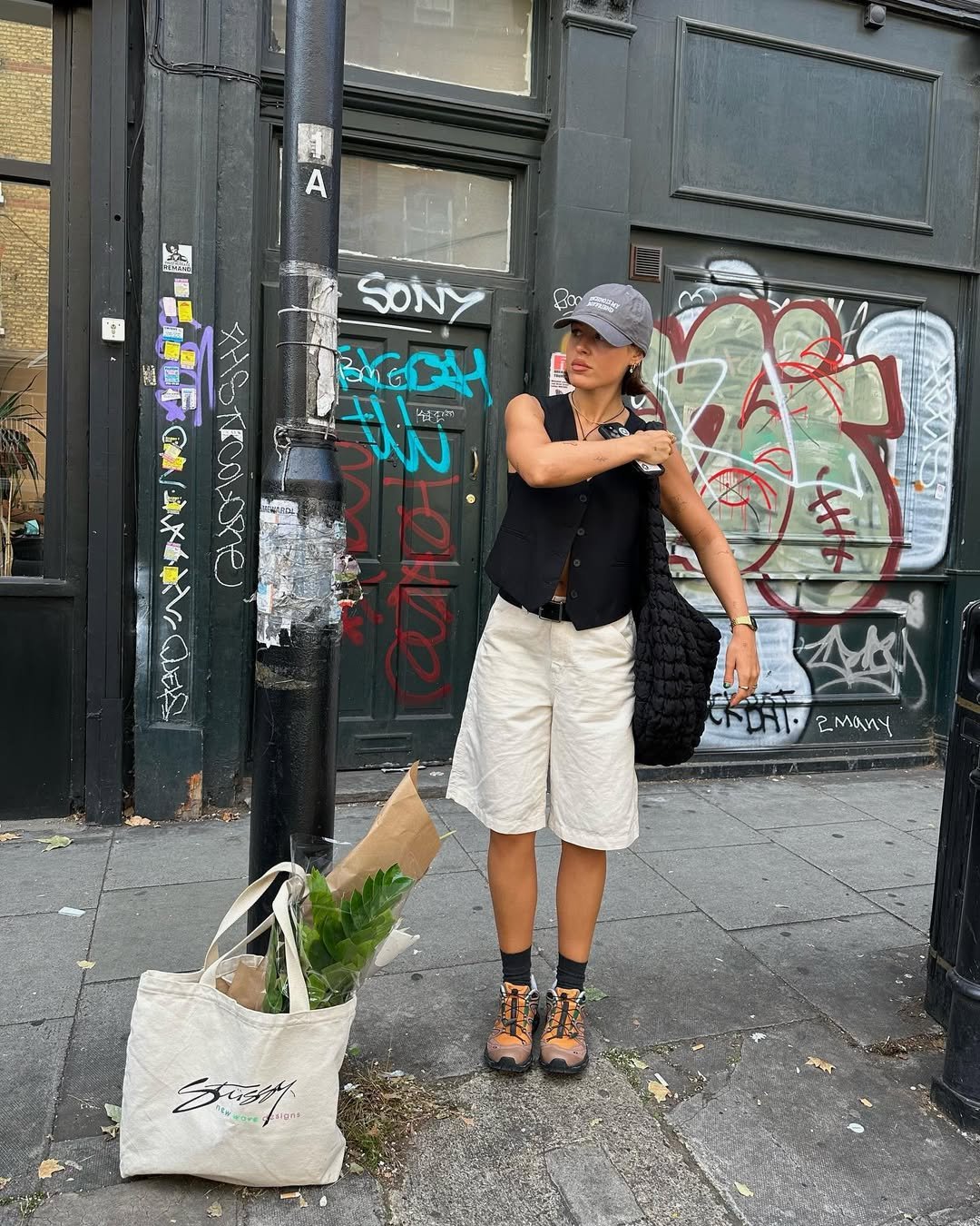 Pessoa usando bermuda branca e tênis laranja, posicionada em frente a parede grafitada. bolsa de compras ao lado.