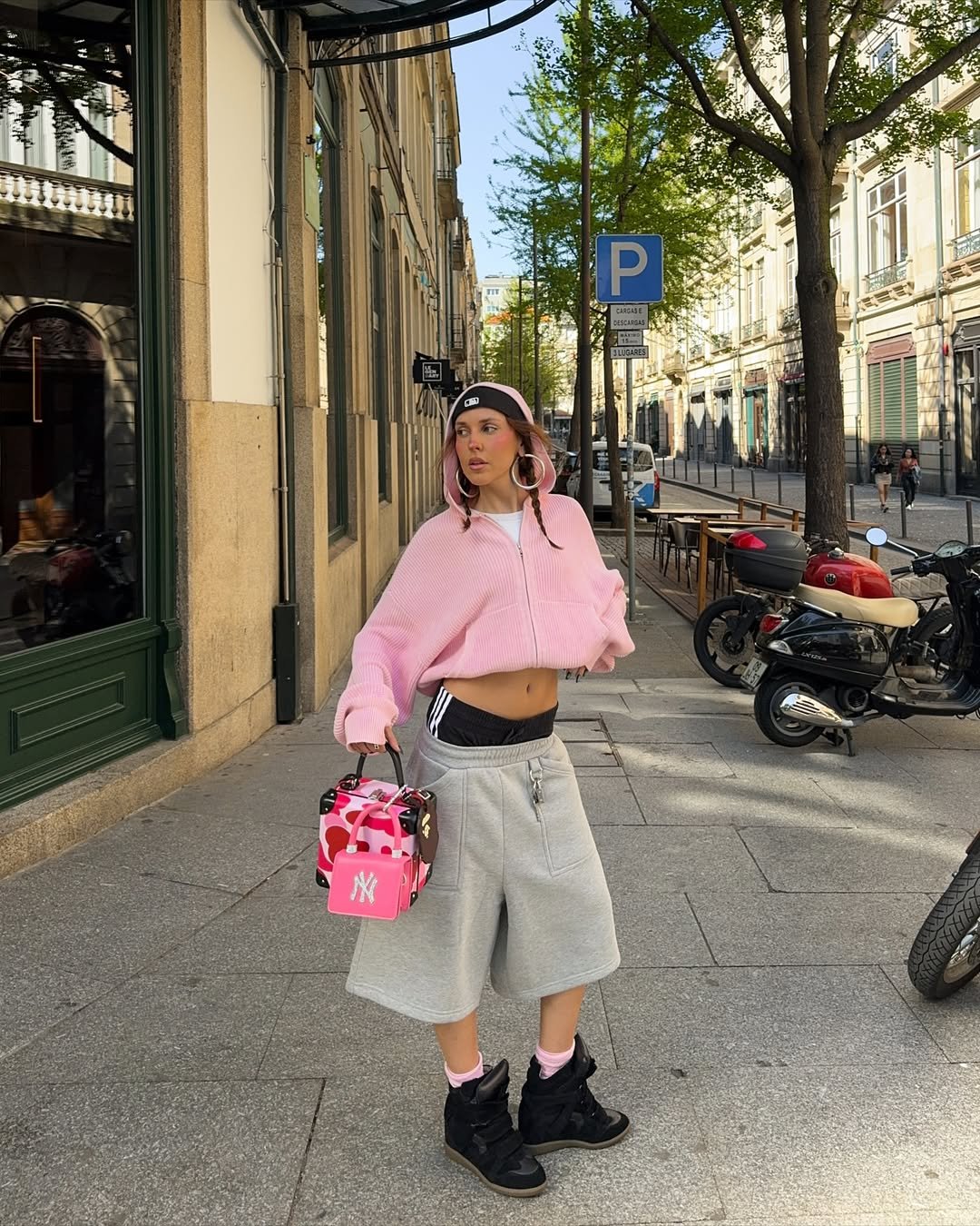 Pessoa usando bermuda cinza e tênis preto, segurando bolsa rosa, em rua movimentada com árvores.