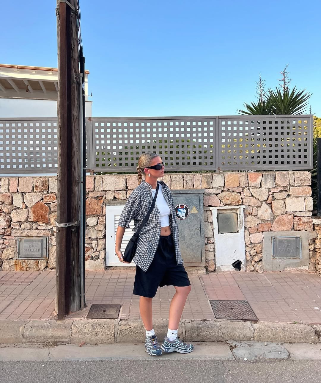 Pessoa com camisa xadrez aberta, bermuda e tênis, posa em frente a um muro de pedras.