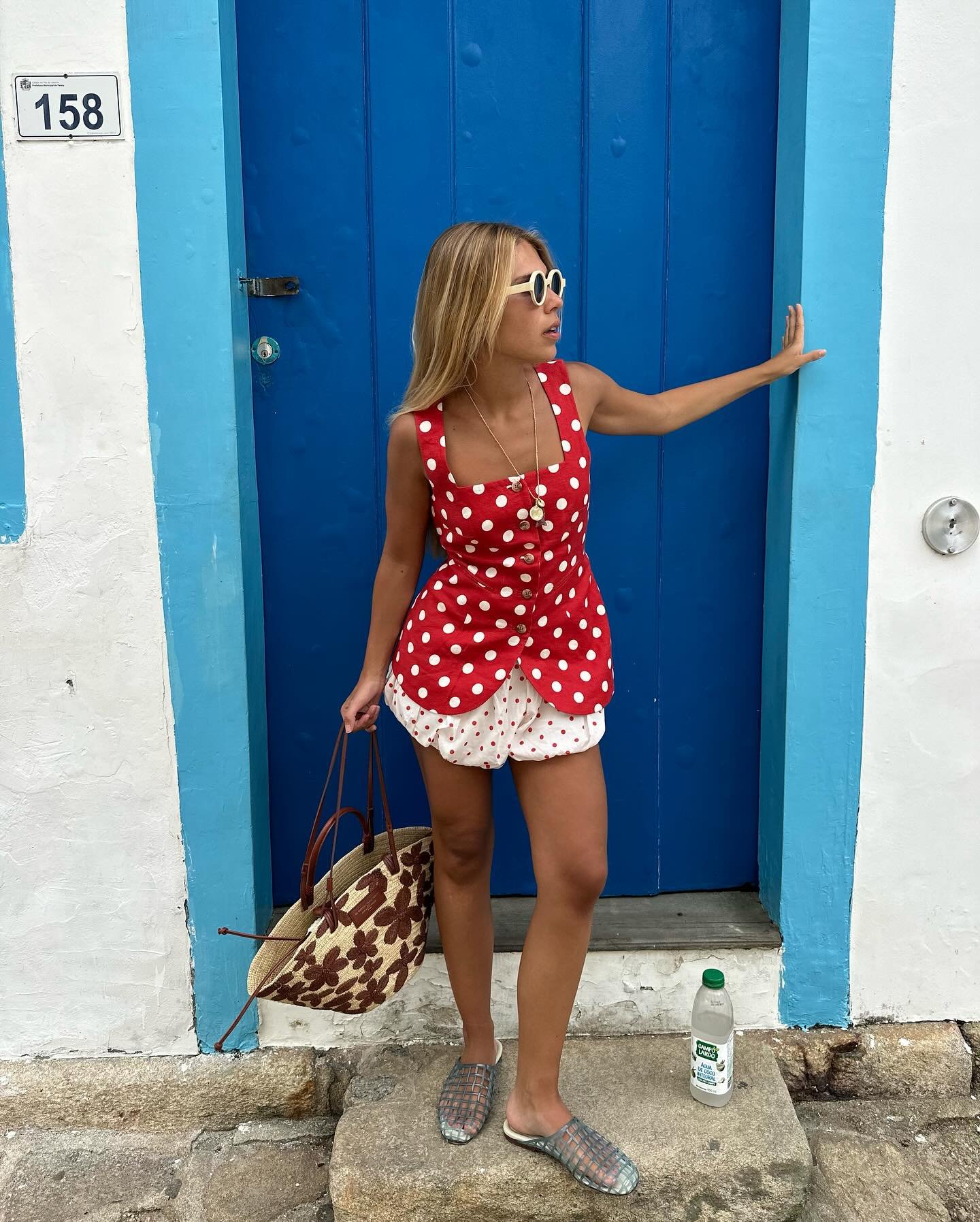 Pessoa com looks com blusa vermelha de bolinhas, óculos escuros e bolsa estampada diante de porta azul vibrante.