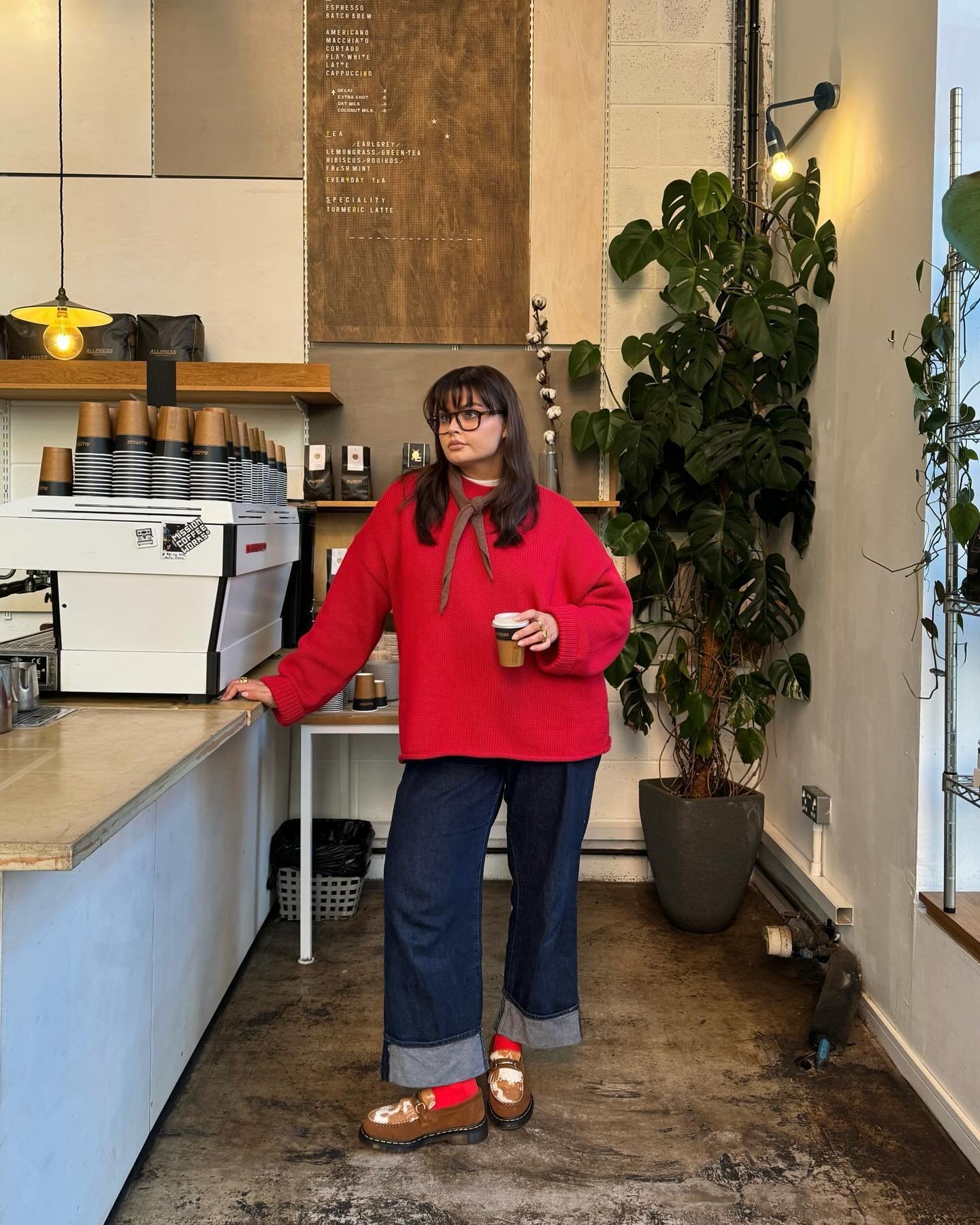 Pessoa em cafeteria com blusa vermelha, calça jeans e sapatos marrons, segurando uma bebida. Looks com blusa vermelha