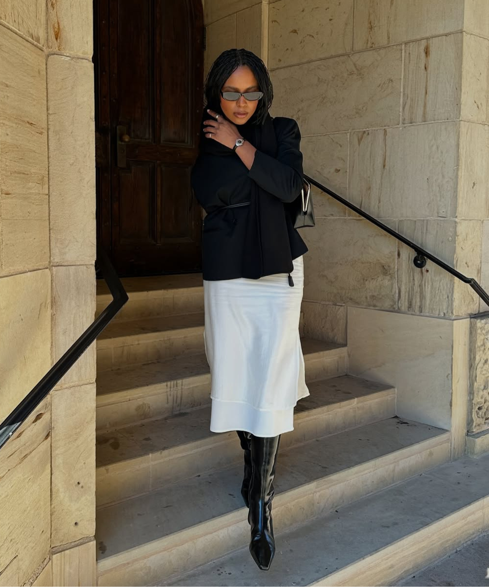 Pessoa em escada com óculos de sol e look elegante, usando bota preta, casaco preto e saia clara.