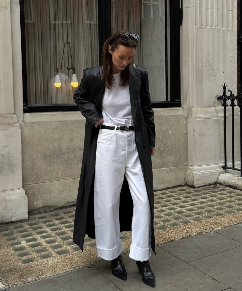 Pessoa usando casaco preto longo, calça branca e botas pretas, em frente a um prédio urbano.
