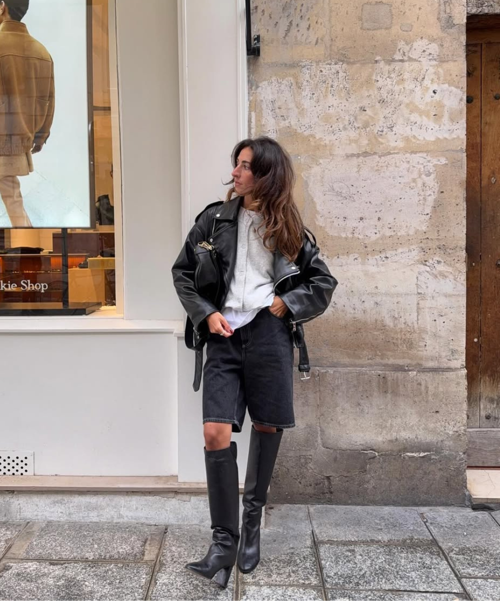 Usuário com jaqueta e bermuda pretas, combinando com botas. Olhando para uma vitrine na rua. Ideia de looks com bota preta.
