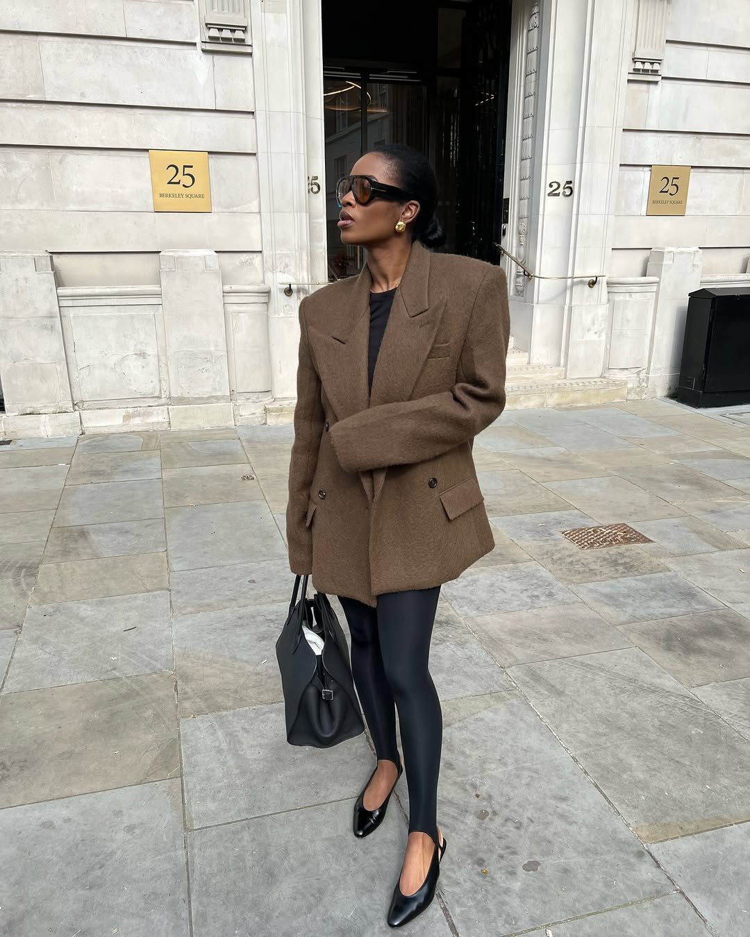 Mulher usando blazer marrom, legging fusô preta e sapatos pretos, segura bolsa. Ao fundo, prédio elegante.