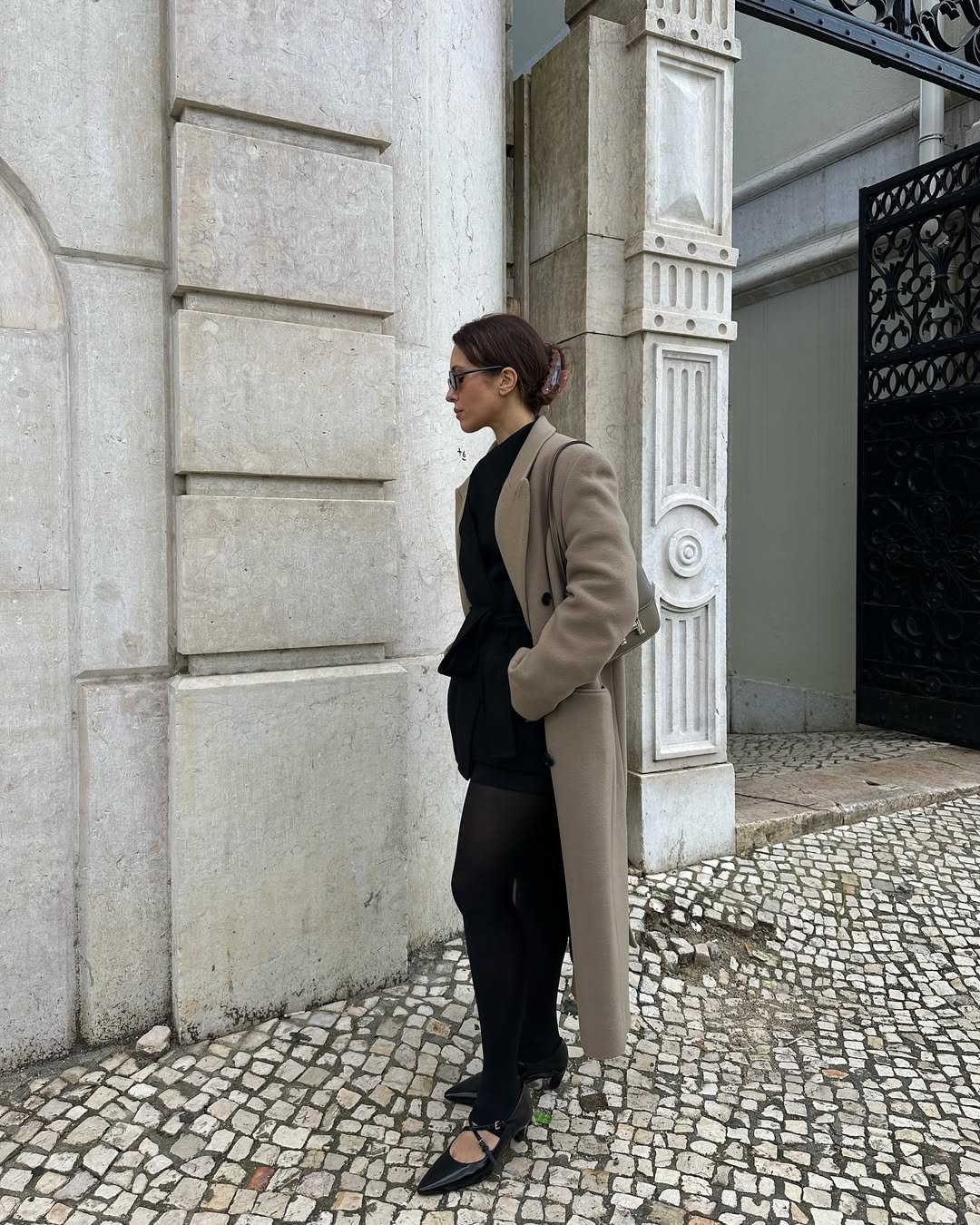 Mulher com casaco bege e legging fusô preta, encostada em parede de pedra em rua pavimentada.