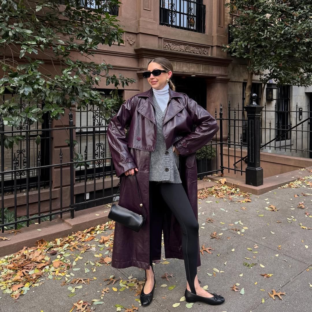 Pessoa usando casaco longo roxo, suéter cinza e legging fusô preta, segura bolsa em rua arborizada.