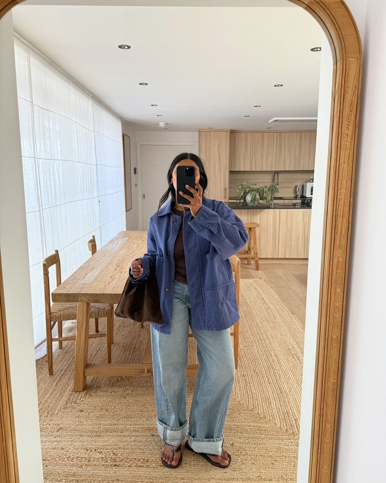 Pessoa em pé em frente a um espelho usando jaqueta azul estilo chore coat, calça preta e tênis branco em sala de estar moderna.