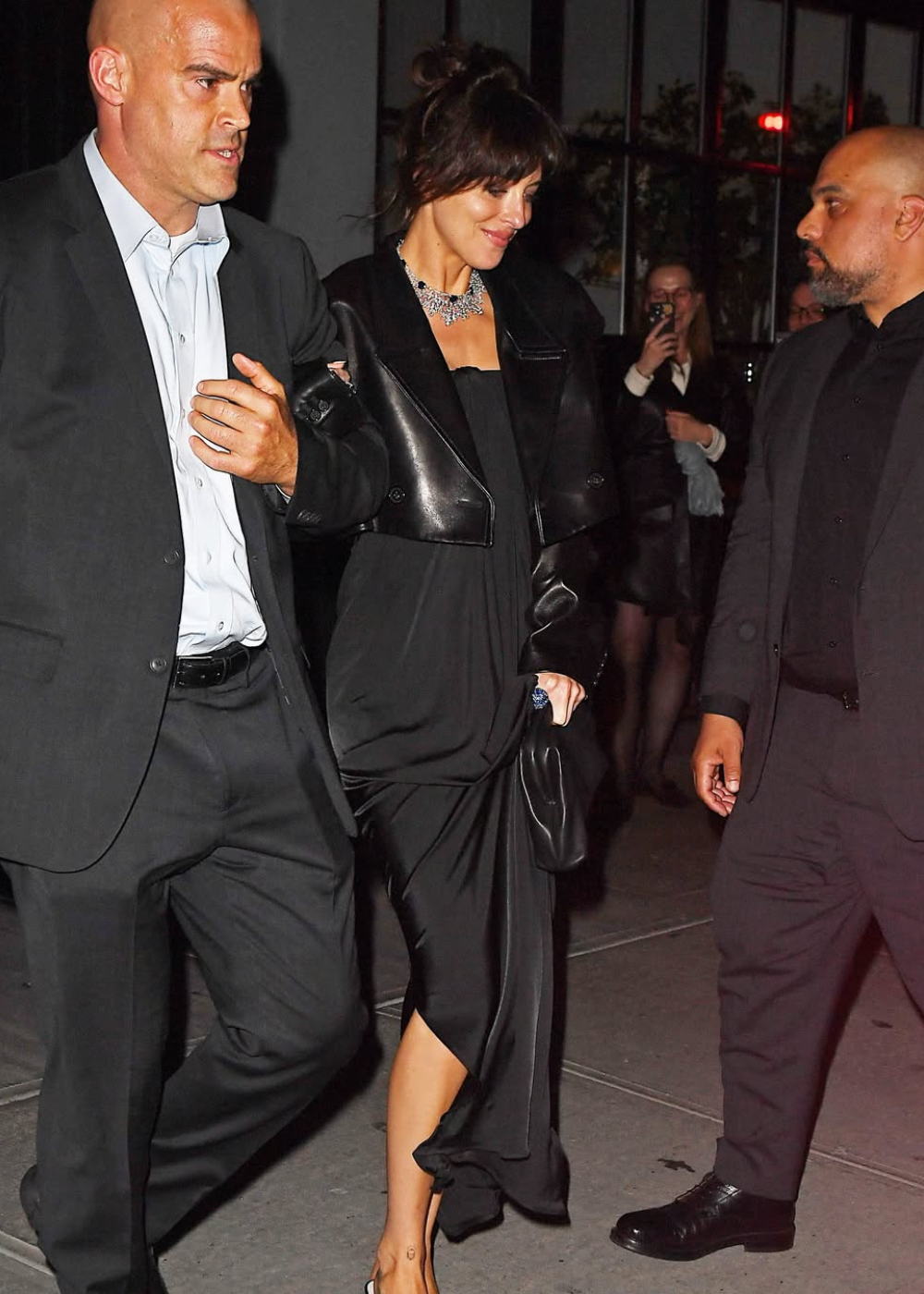 Pessoa em vestido preto elegante, acompanhada por dois homens em ternos. Look sofisticado com um colar brilhante.