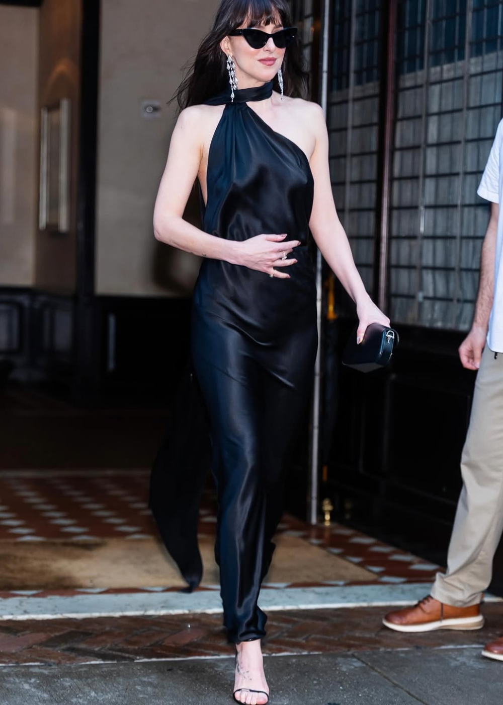 Vestido preto elegante com alça cruzada, brincos longos e óculos escuros. Looks da Dakota Johnson.