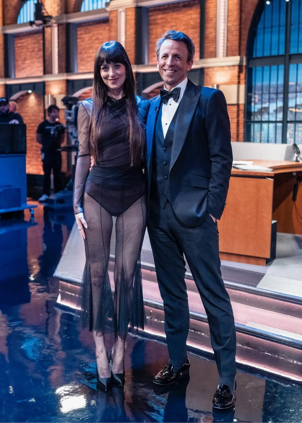 Uma mulher em vestido preto translúcido e um homem de smoking posam sorrindo em um estúdio iluminado.
