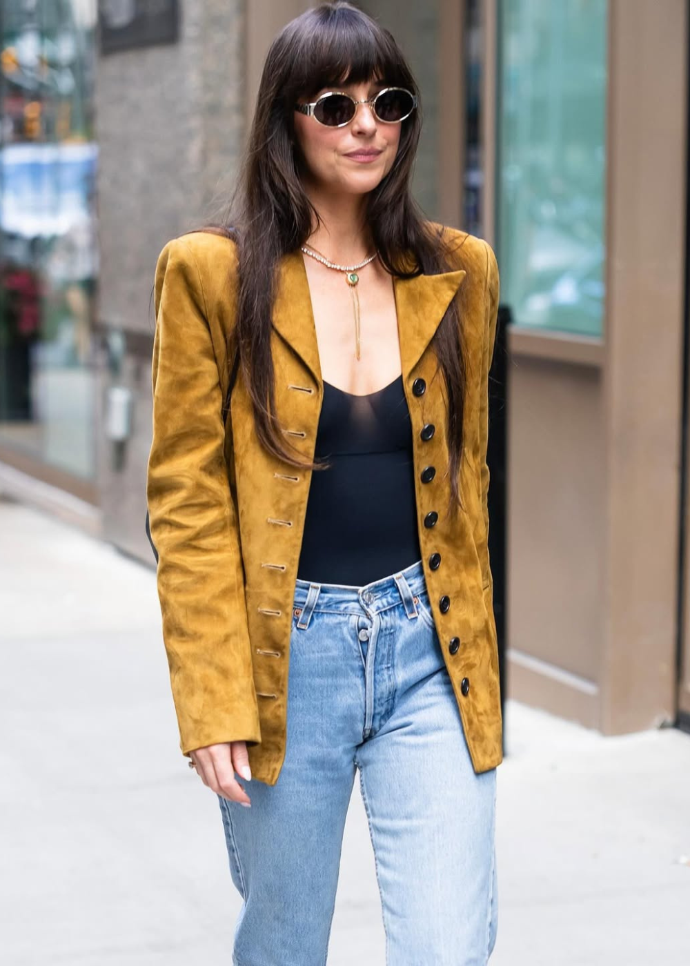 Pessoa com óculos escuros, blazer mostarda e calça jeans clara, caminhando em ambiente urbano. Looks da Dakota Johnson.