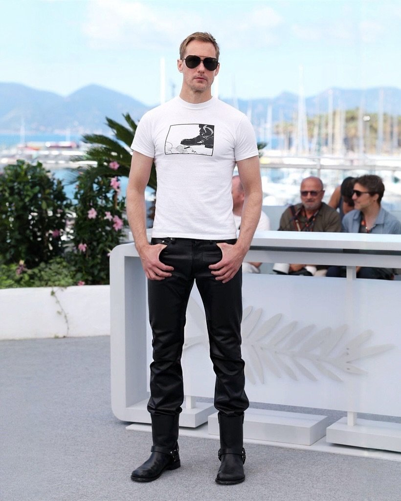 Alexander Skarsgård de camisa branca com estampa em preto e branco em fundo azul.