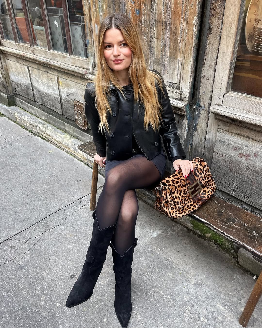 Mulher com casaco preto, botas e bolsa de estampa animal, sentada em banco de madeira desgastado.