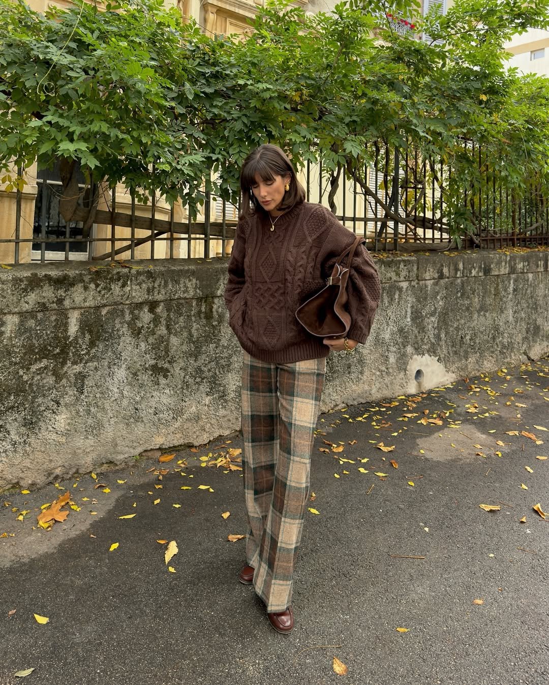 Pessoa usa suéter marrom e calça xadrez, idealizando looks para Campos do Jordão. Caminha em calçada, folhas ao chão.