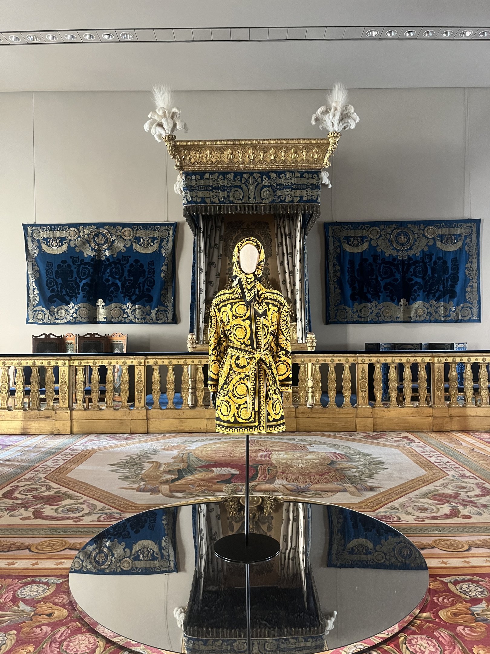 Manequim com traje dourado e preto em ambiente luxuoso, destacando estilo Louvre Couture. Fundo com tapetes ricos e detalhes dourados.