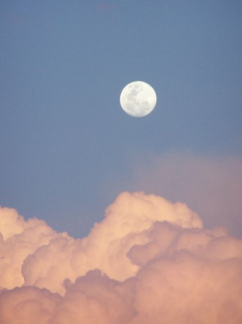 Lua cheia no céu azul, sobre nuvens rosadas ao entardecer.