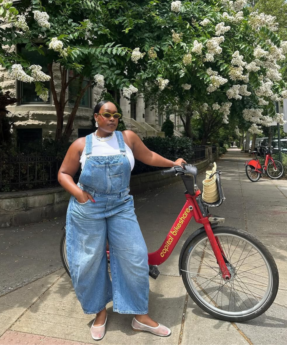 Pessoa com macacão jeans e óculos de sol apoia-se em bicicleta vermelha em calçada arborizada.