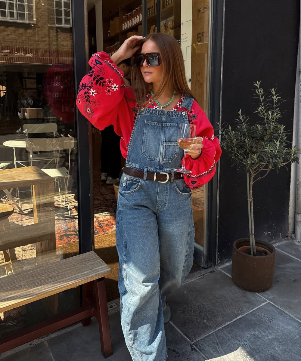 Mulher com blusa vermelha bordada e macacão jeans, segura uma taça de vinho. Ela usa óculos escuros.