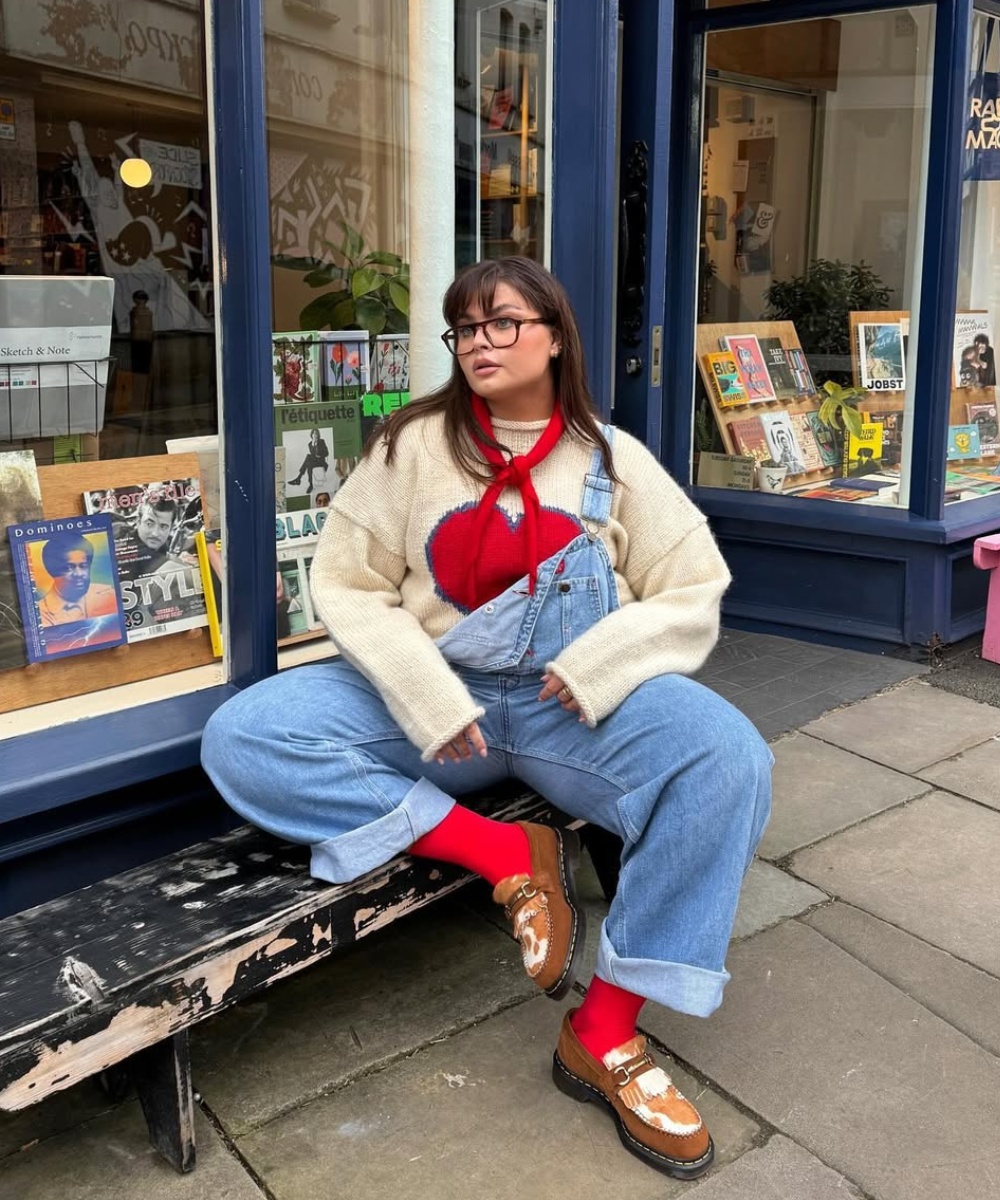 Pessoa sentada usa macacão jeans com suéter de coração vermelho e sapatos marrons, em frente a uma loja de livros.