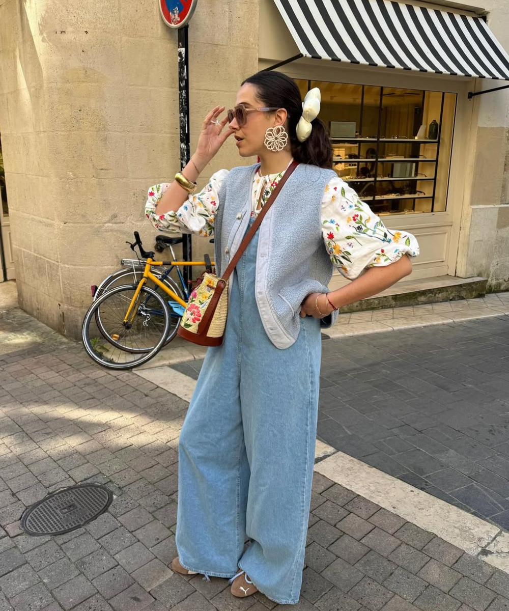 Mulher com blusa floral e calça jeans larga, usa grandes brincos e segura óculos de sol. Bicicleta amarela ao fundo.