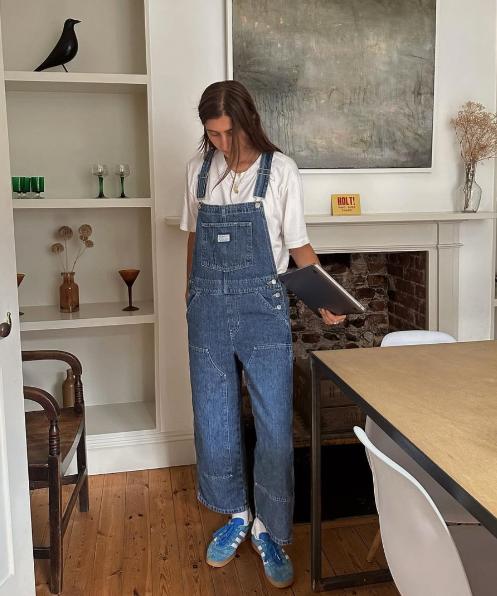 Pessoa em macacão jeans azul, segurando um laptop em uma sala com decoração minimalista.