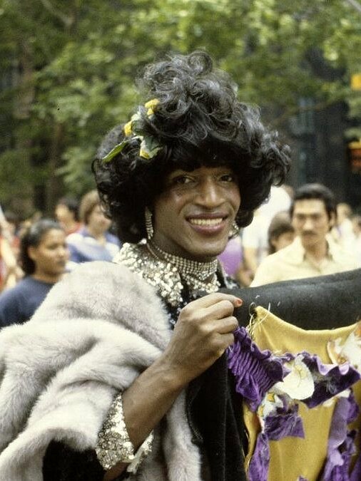 Pessoa sorridente em evento, vestindo roupa colorida e acessórios, celebrando diversidade e orgulho LGBTQIAPN+.