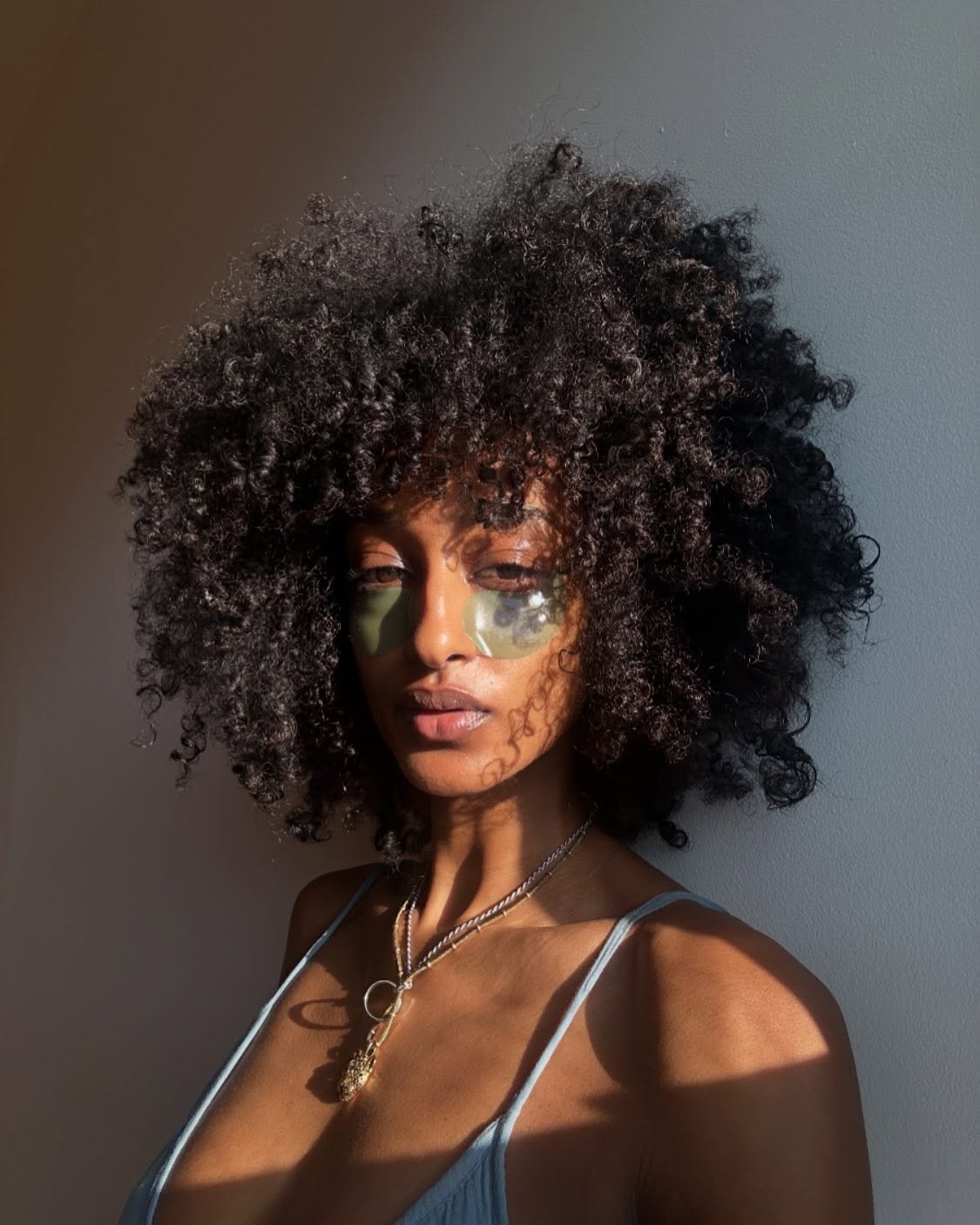 Mulher com cabelo cacheado e máscara facial sob os olhos, destacada pela luz natural, como um protetor de pele e usando protetor térmico.