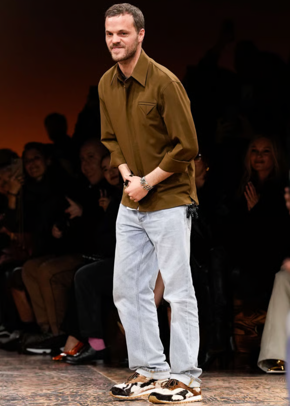 Pessoas observam desfile de moda com foco em um designer sorrindo no centro da passarela.