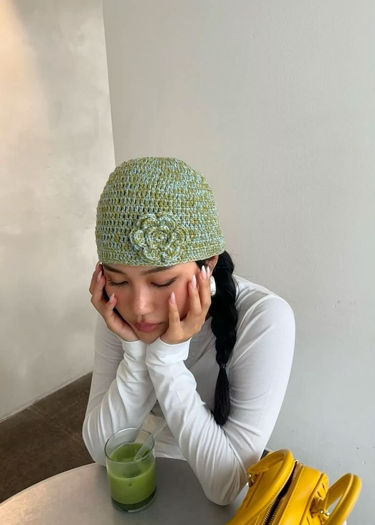 Pessoa com chapéu de crochê verde e camiseta branca apoia a cabeça nas mãos, ao lado de um suco verde. A mesma