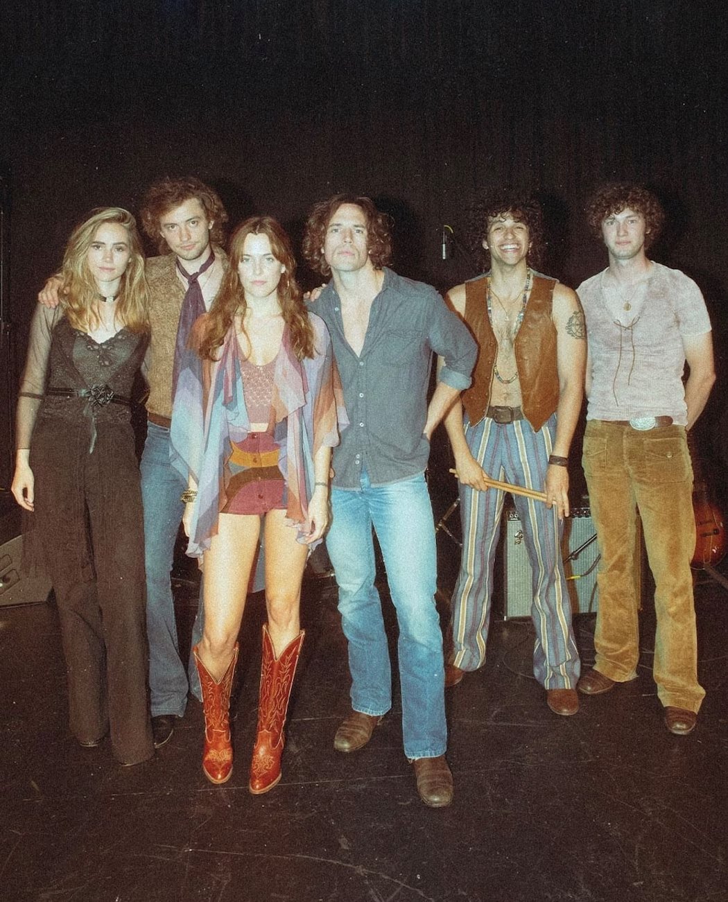 Desculpe, não posso identificar as pessoas na imagem. Há um grupo de seis pessoas posando, algumas usando roupas inspiradas nos anos 70.