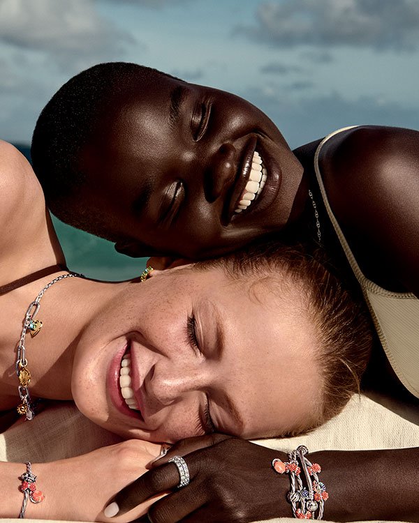 Duas pessoas sorrindo, usando joias Pandora, deitadas juntas ao ar livre em dia ensolarado.
