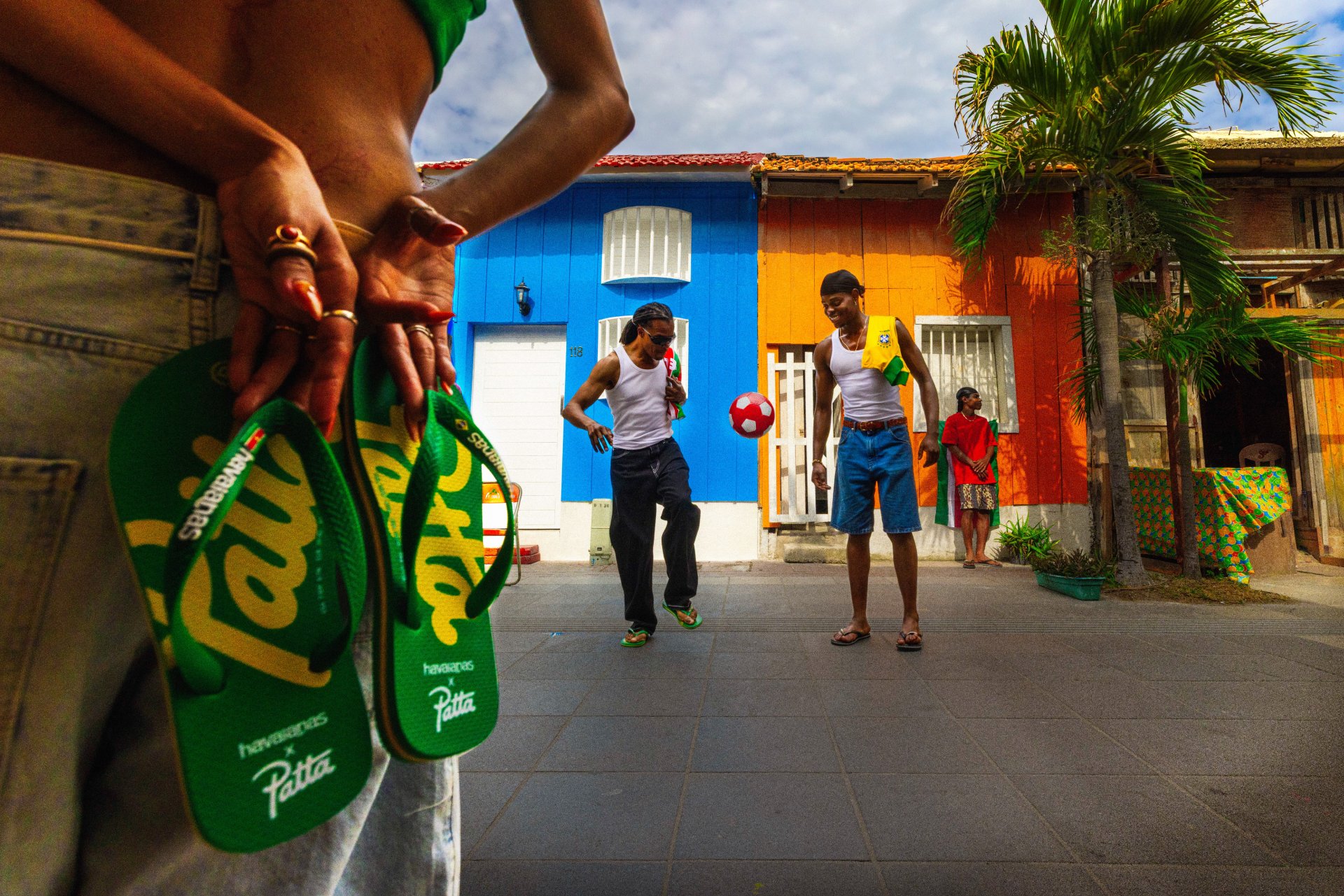Pessoas jogam futebol de rua em frente a casas coloridas. Chinelo verde da Patta em destaque.