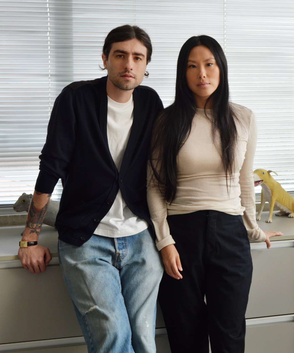 Duas pessoas de pé em frente a uma janela com persianas, cercadas por objetos decorativos. Pedro e Paula, diretores criativos da P. Andrade.