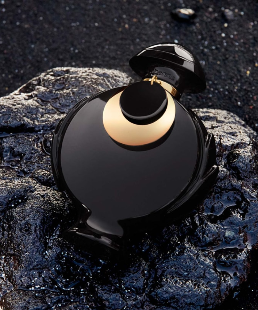Frasco escuro de perfume com detalhes dourados, repousando sobre rochas pretas.